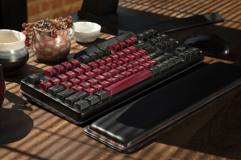 GMK Red Samurai Custom Keycaps on a black keyboard on a wooden desk with a black leather wrist pad with the sun shinning through the blinds