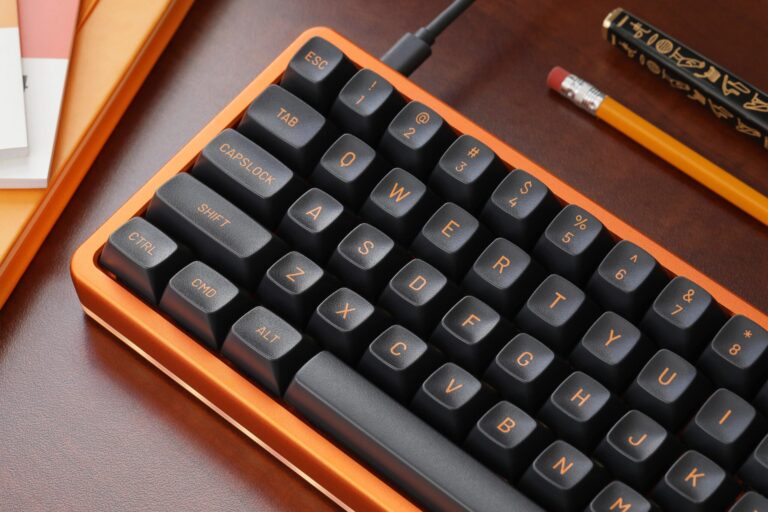 Orange keyboard with black keycaps with orange lettering on a wooden desk with an orange pencil